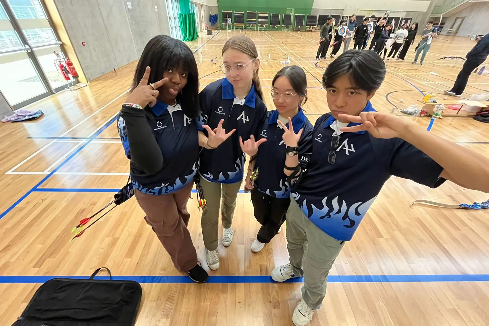 Four student archers in matching club shirts posing in a sports hall at an ISAA competition, with the shooting line behind them